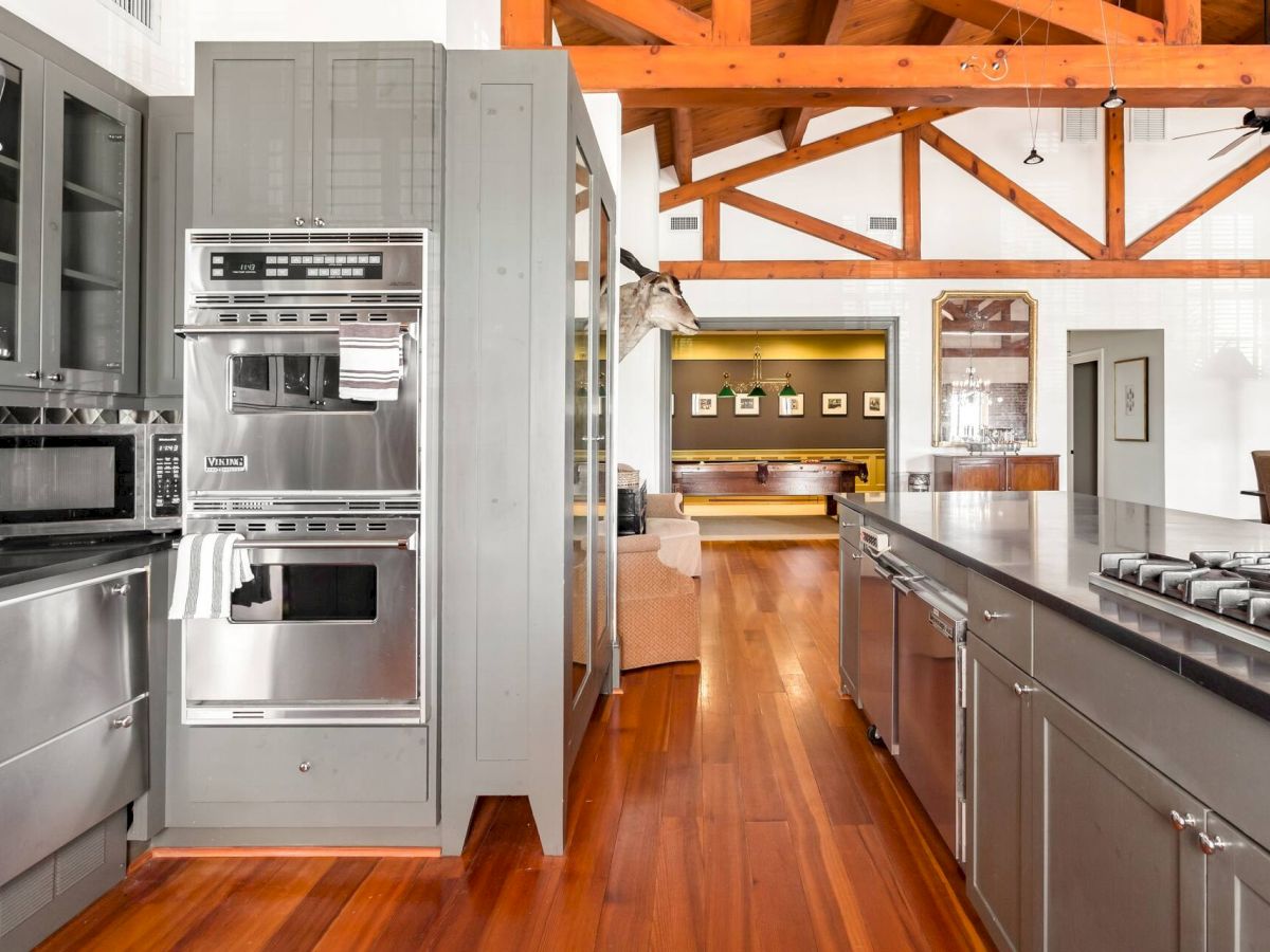 A modern stainless steel kitchen with a double wall oven, large refrigerator, island prep area, sleek cabinetry, and warm wood floors under exposed wooden beams.