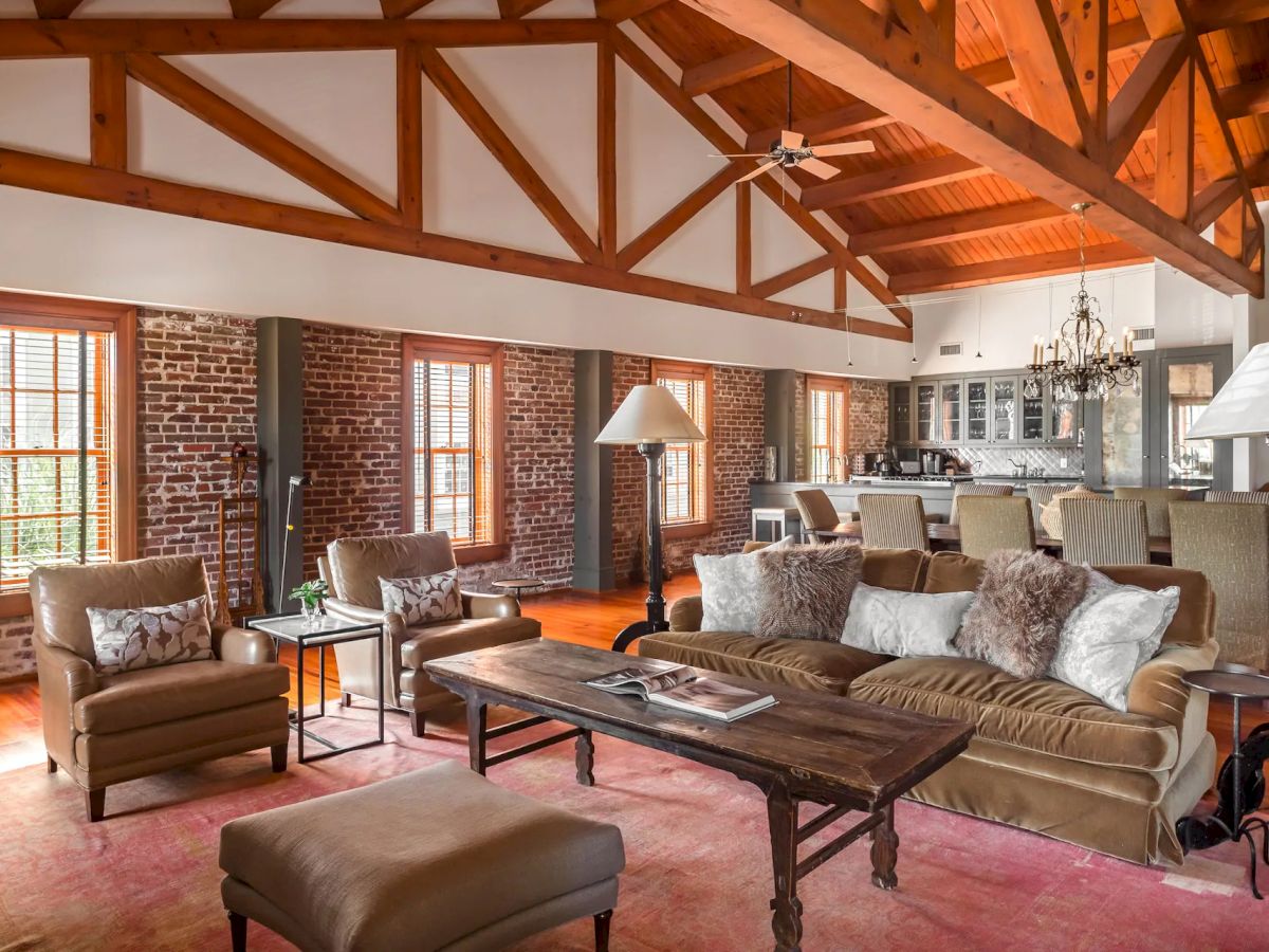 A spacious, rustic living room with exposed wooden beams, brick walls, large windows, and cozy seating including sofas, chairs, and a coffee table, plus warm lighting.