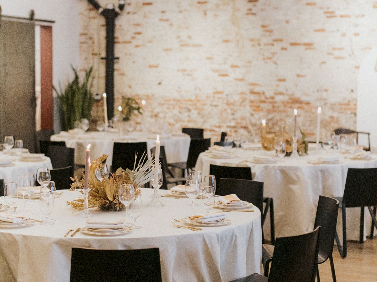 A stylish dining room set for a formal event: round tables with white tablecloths, black chairs, elegant table settings, warm lighting, and a brick accent wall.
