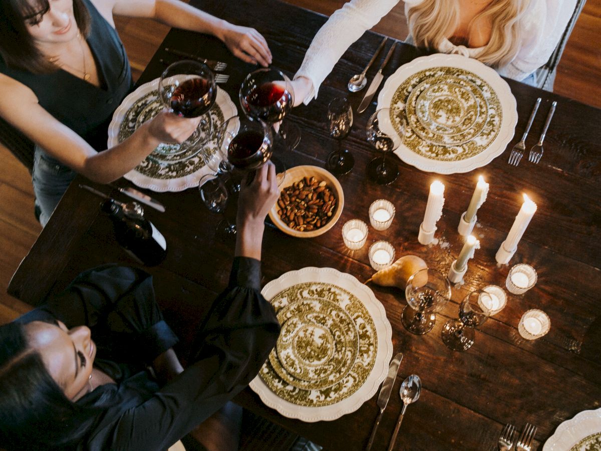 A group of seven people sit around a candlelit dinner table, toasting with glasses, plates, bowls of nuts, and a warm, intimate meal atmosphere.
