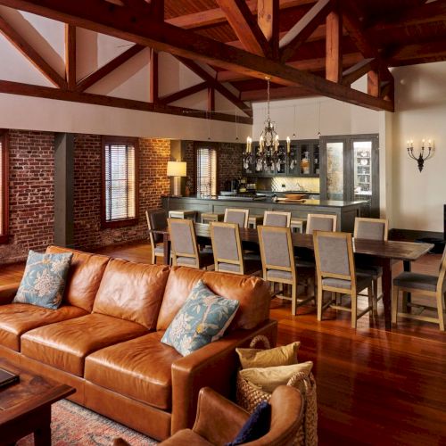 A cozy loft interior with exposed wooden beams, a brick wall, leather sofa, dining table, and a modern kitchen bar.