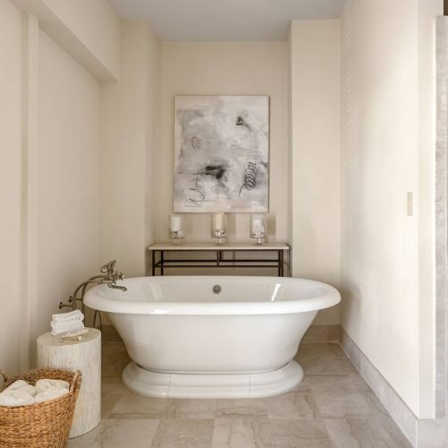 A bright bathroom with a freestanding oval tub, wicker baskets, a wall mirror, and a framed artwork above a small console; shower on the right.