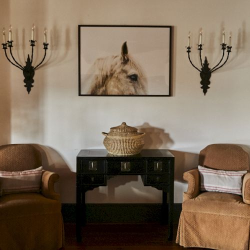 Two beige armchairs flank a dark console with a woven basket on top, a framed horse photo above, and wall sconces on either side.