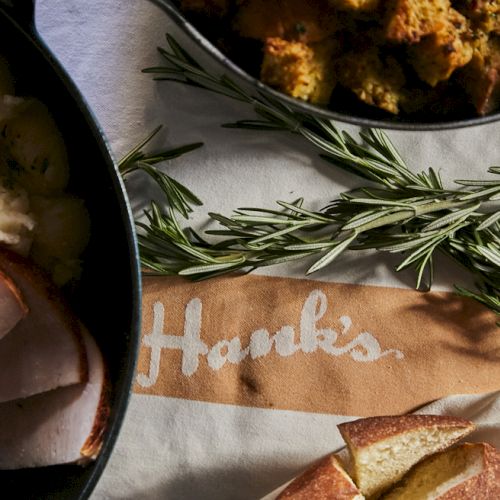 A skillet with sliced roasted meat, rosemary sprigs, and a napkin reading &ldquo;Thanks,&rdquo; plus crusty bread and fried bits nearby, all on a table.