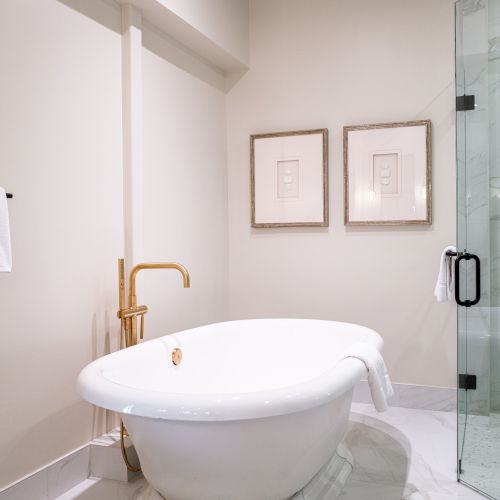 A modern bathroom with a white oval sink, gold faucet, framed wall art, and a glass shower beside a towel.