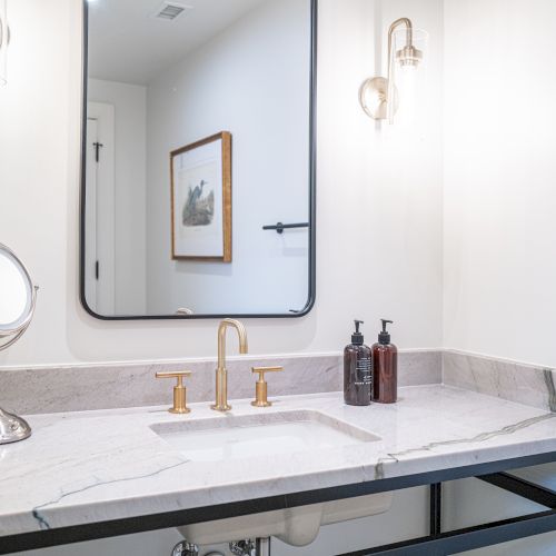 A modern bathroom vanity with a marble sink, gold faucets, soap bottles, and wall sconces reflecting in a mirror.