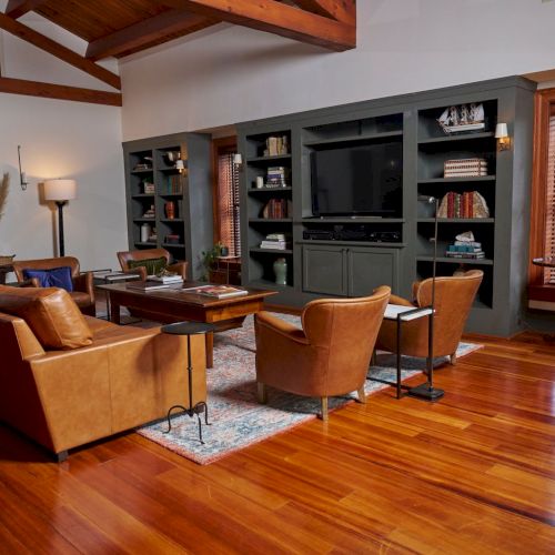 A cozy living room with a leather sofa, armchairs, built-in TV shelving, warm wood floors, and a modern lamp under exposed beams.
