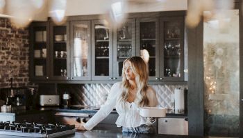 A woman in a bright kitchen, standing at a marble island with cabinets behind, warm lights creating a cozy, slightly blurred foreground.