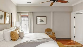 A bright hotel-style bedroom with a large bed, white linens and tan pillows, a ceiling fan, a chair, artwork, and a doorway to the right.