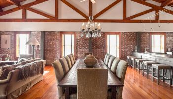 A spacious dining area with a long wooden table, upholstered chairs, brick walls, large windows, and a chandelier, plus a living area nearby.