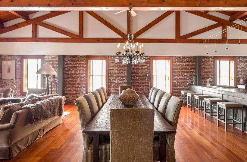 A spacious dining area with a long wooden table, upholstered chairs, brick walls, large windows, and a chandelier, plus a living area nearby.