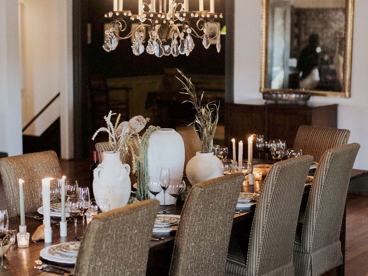 A formal dining room with a lit chandelier, tall candles, glassware, floral decor, and upholstered chairs around a set table.